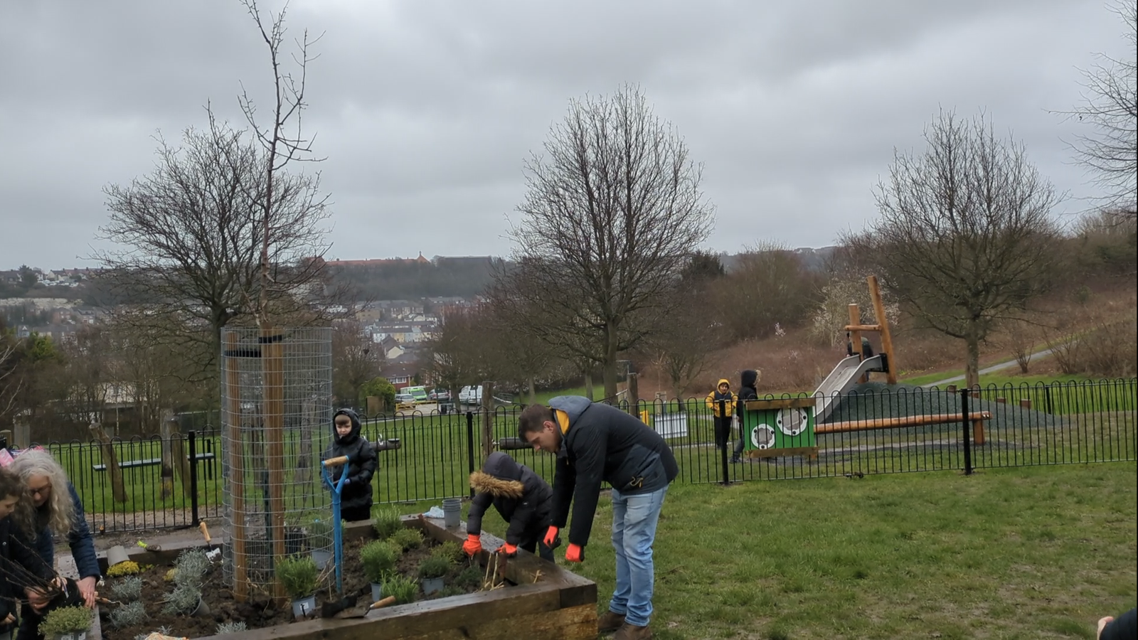 A picture of people planting plants in a shrub bed. More play equipment can be seen in the background, including a slide and balance beam.