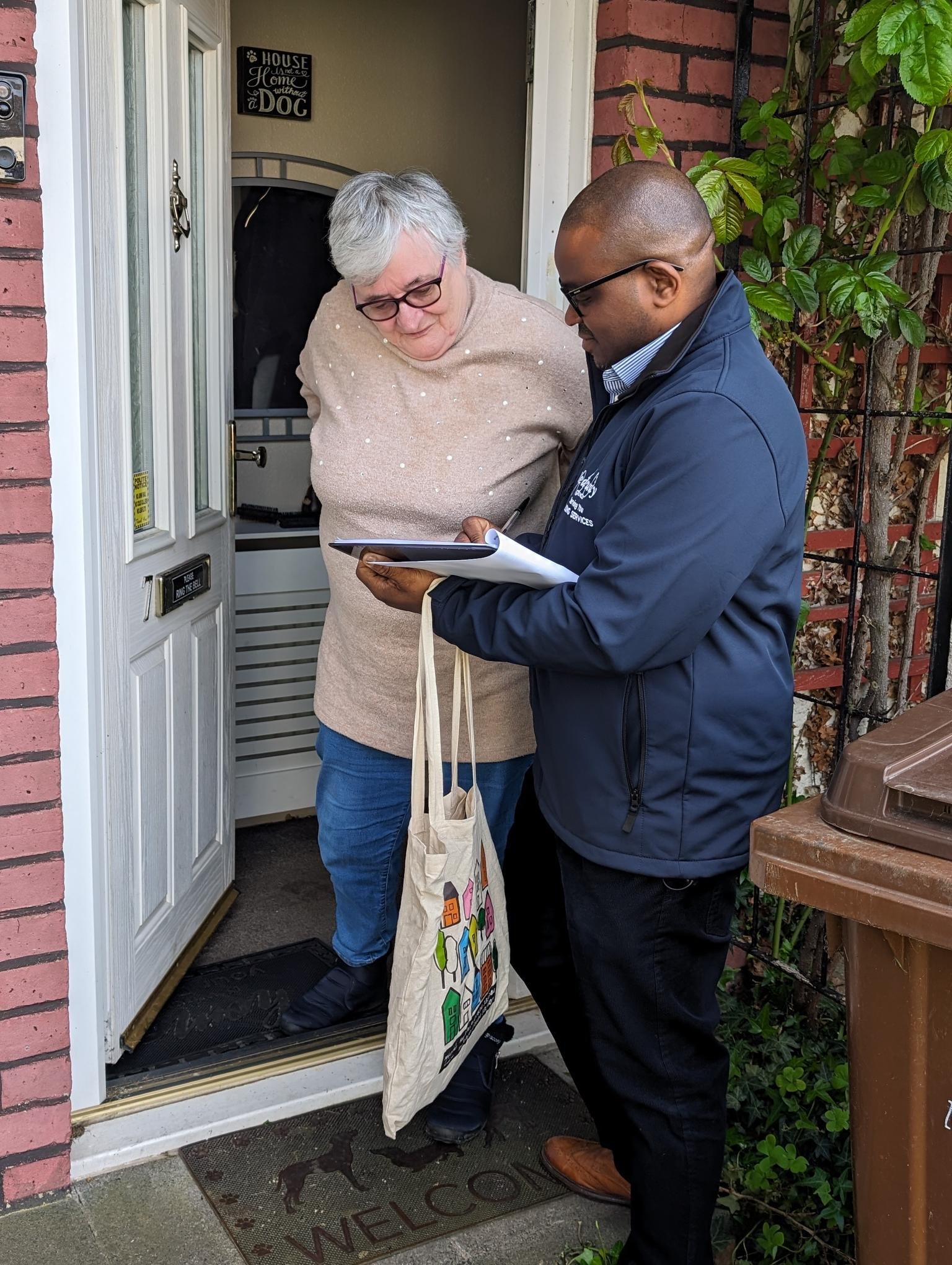 A man is stood talking to a woman in front of her house with the door open. He is holding a clipboard in his hand, and she is looking down at it as well.
